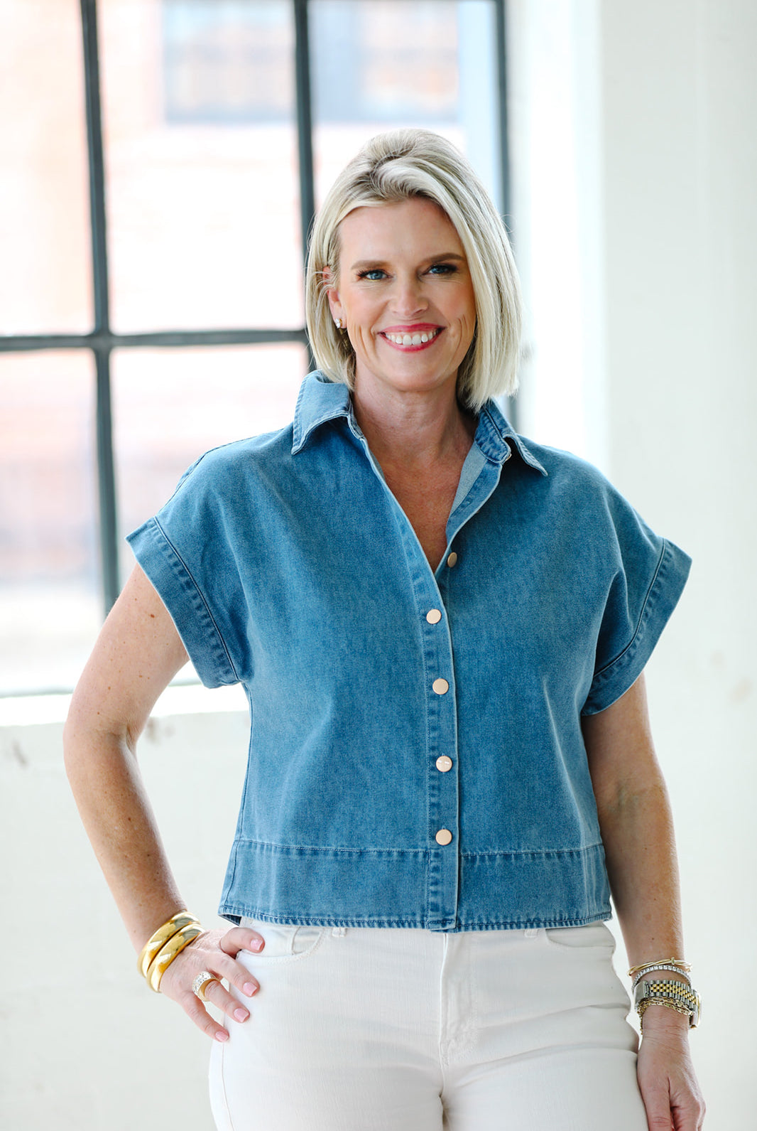 Woman wearing a denim shirt standing in a bright room with large windows.