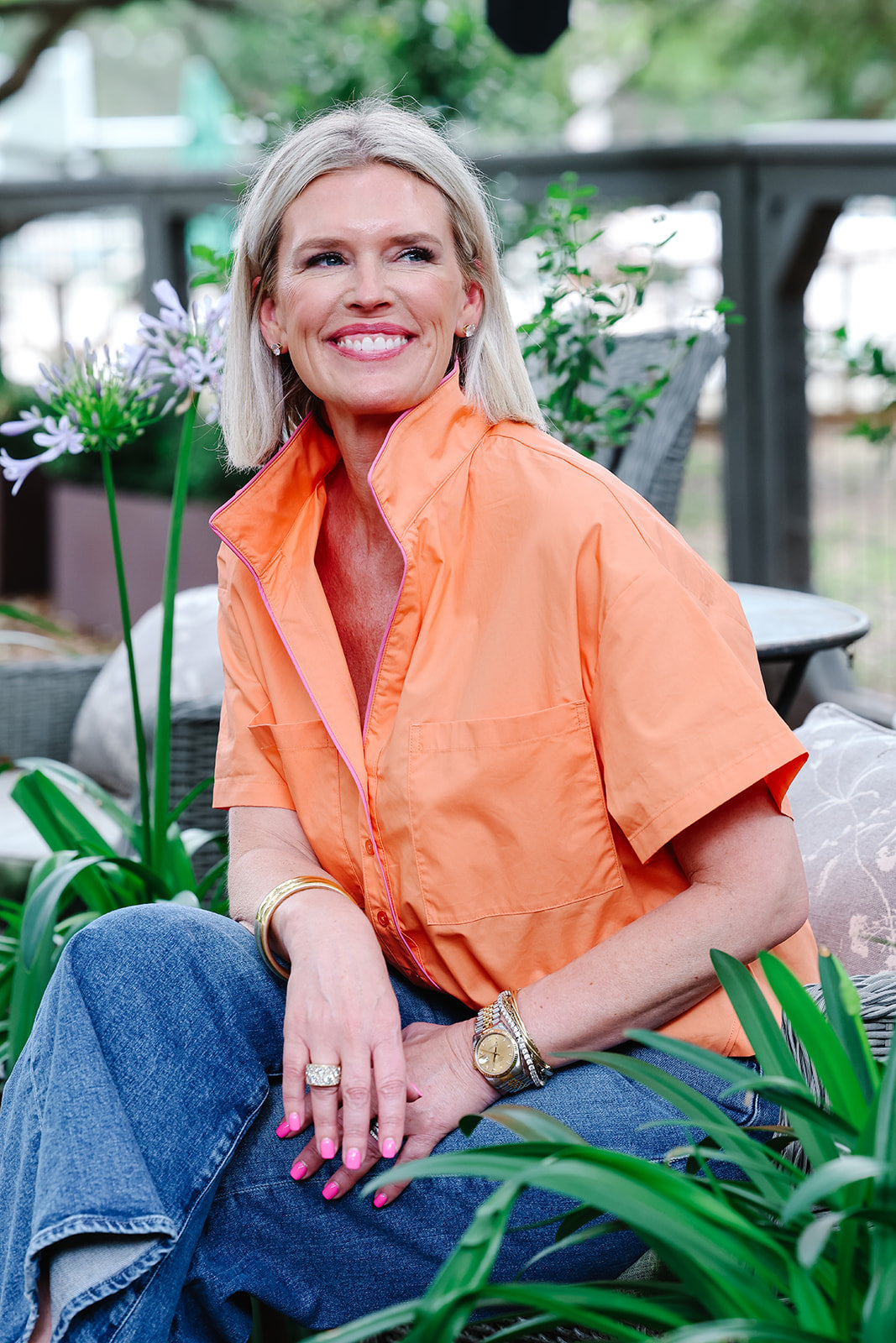 Woman wearing an orange shirt and blue jeans sitting among plants.