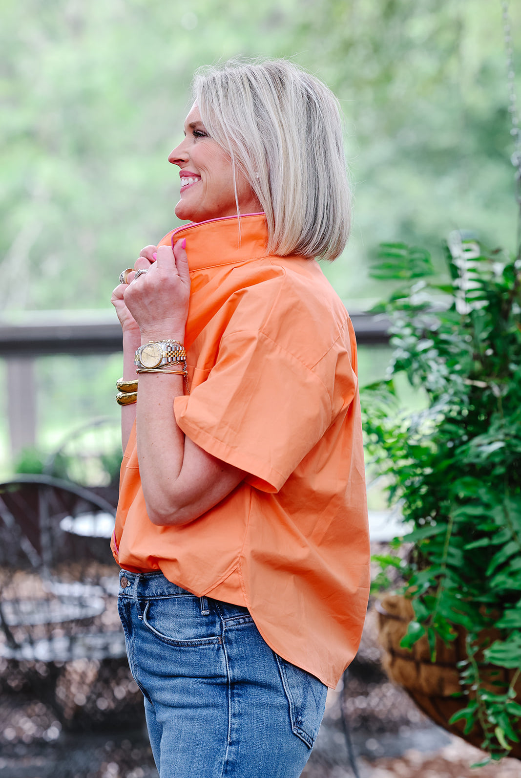 Woman wearing an orange shirt and blue jeans standing outdoors with greenery in the background