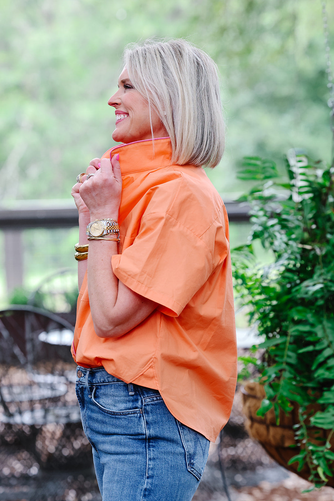 Woman wearing an orange shirt and blue jeans standing outdoors with greenery in the background