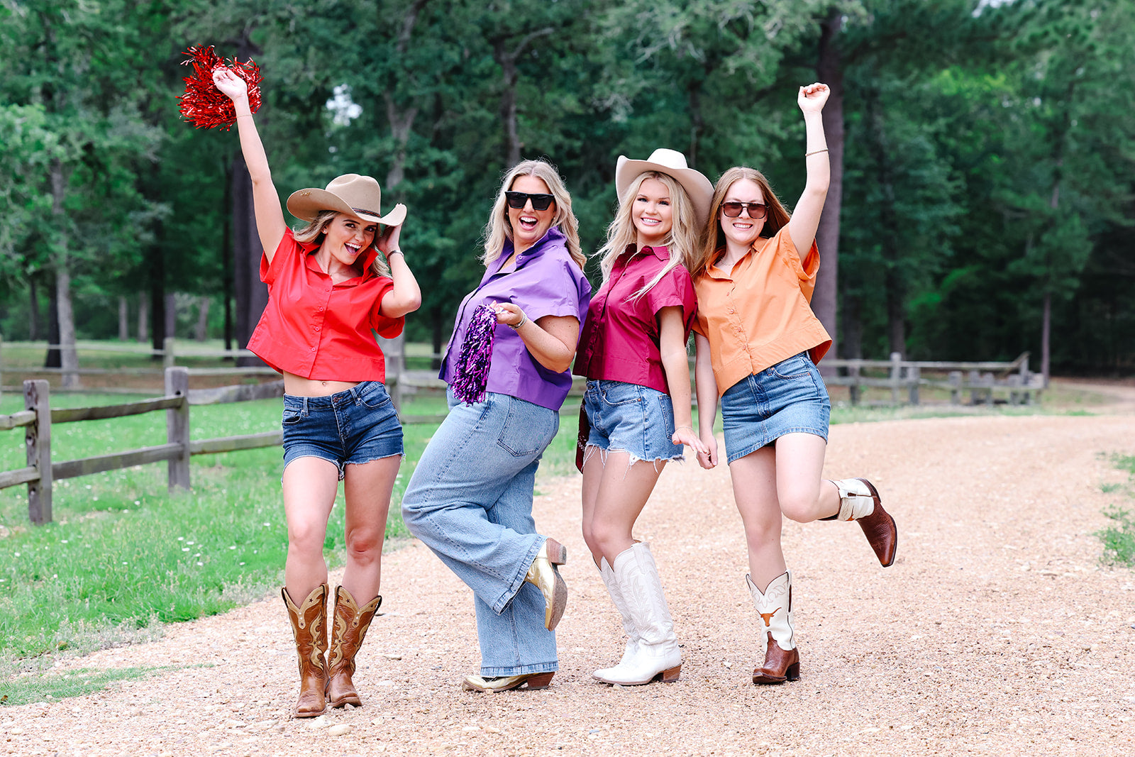 Four women in cowboy hats and boots posing on a dirt path with trees in the background