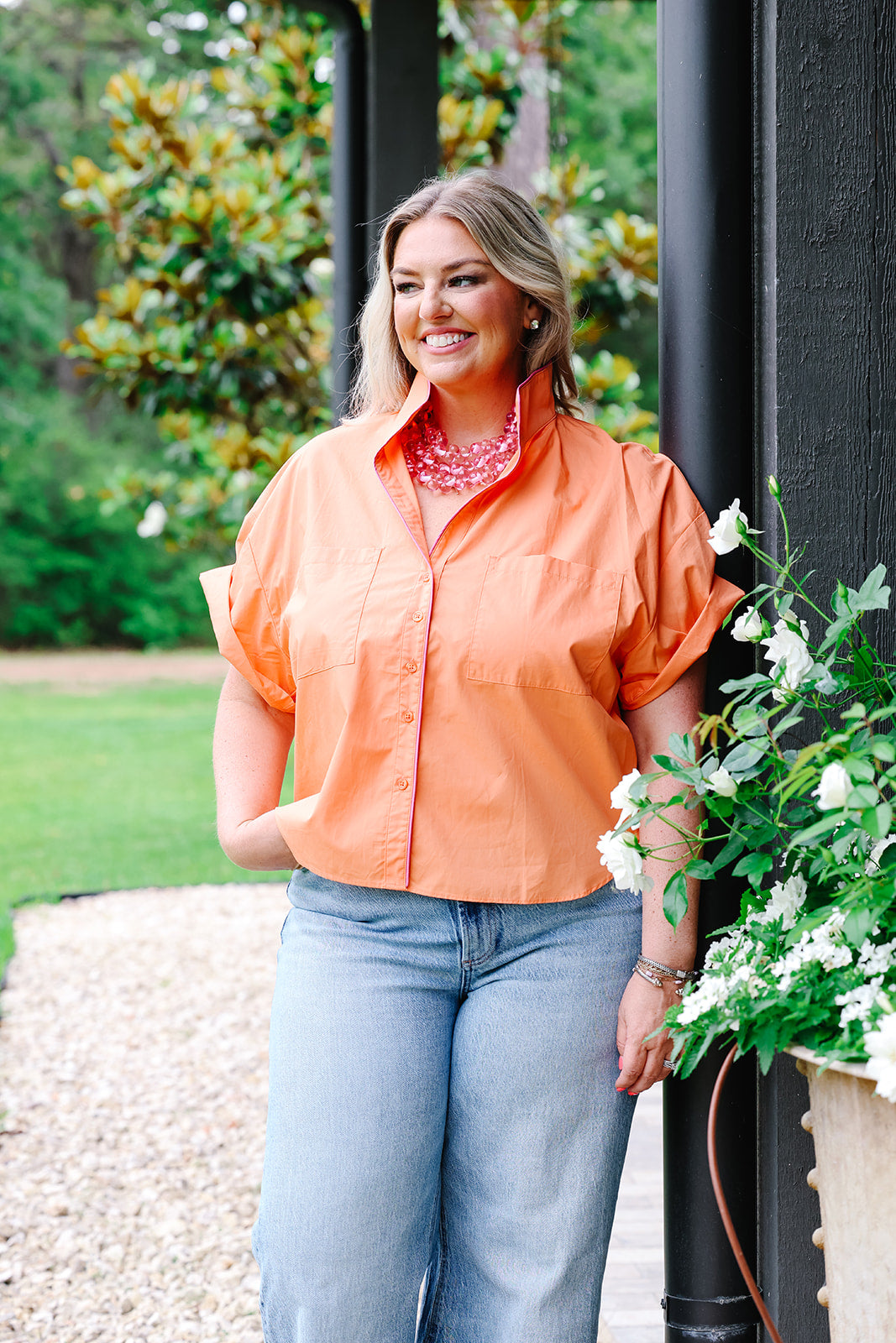 Woman in an orange shirt and blue jeans standing outdoors with greenery in the background