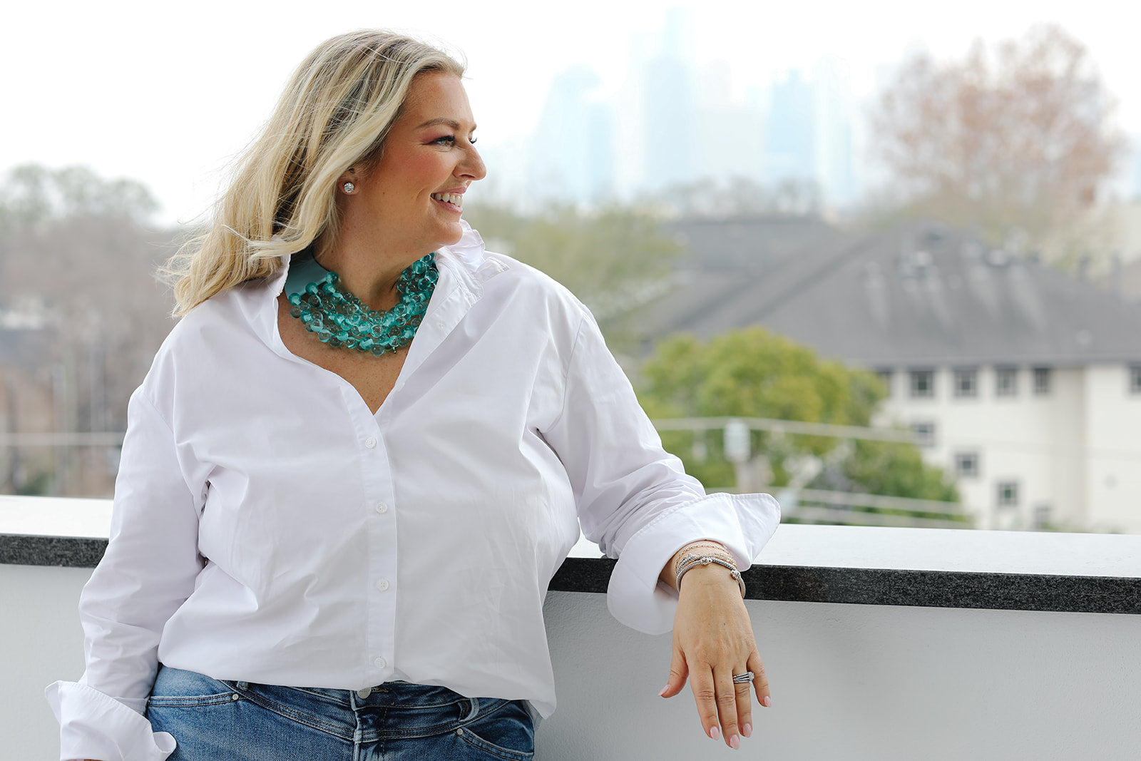 Woman in a white shirt and blue jeans standing on a balcony with a cityscape in the background