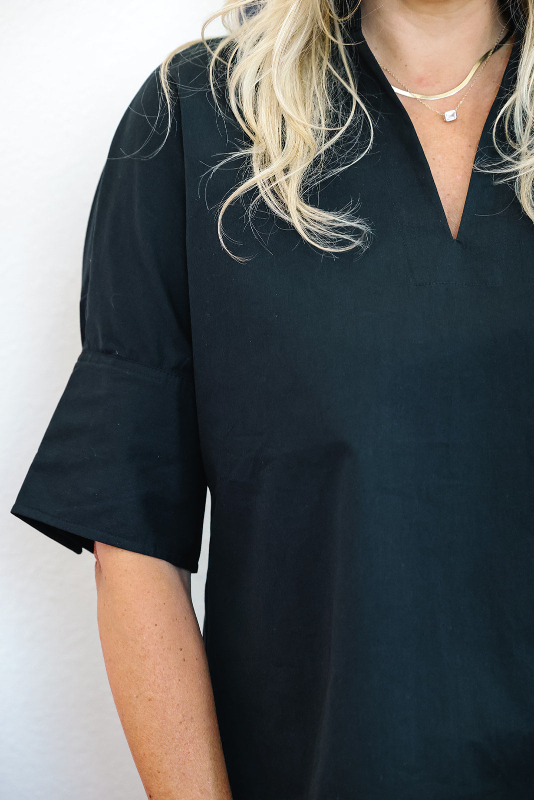 A close-up of a person wearing a black short-sleeve top with a V-neckline and a small chest pocket, paired with layered gold necklaces against a plain light background.
