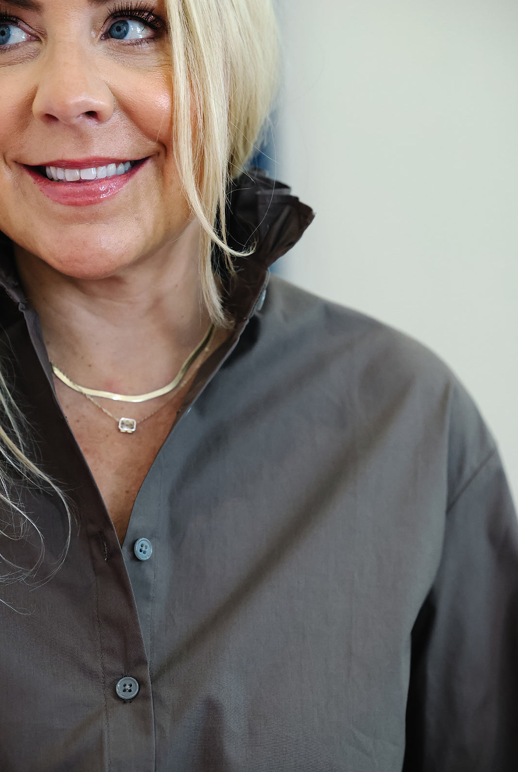 A close-up lifestyle shot of a smiling blonde woman wearing a dark brown button-down shirt and a delicate gold chain necklace with a small square pendant.