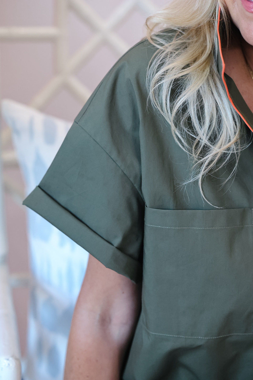 A close-up of a person wearing an olive green scrub top, showing the chest pocket and rolled-up sleeve, with blonde hair and an orange earbud cord visible.