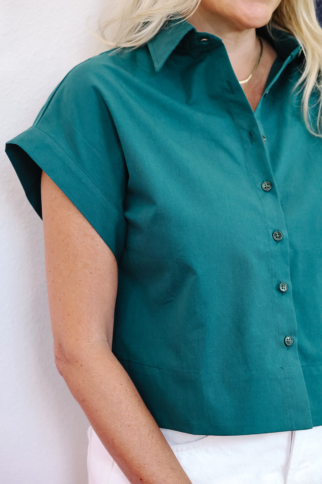 A close-up of a woman wearing a teal green short-sleeve button-up shirt with white pants, shown from the chest down against a plain light background.