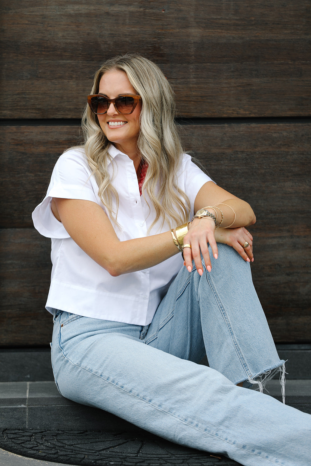 Woman wearing sunglasses and casual white short sleeve blouse sitting against a wooden wall.