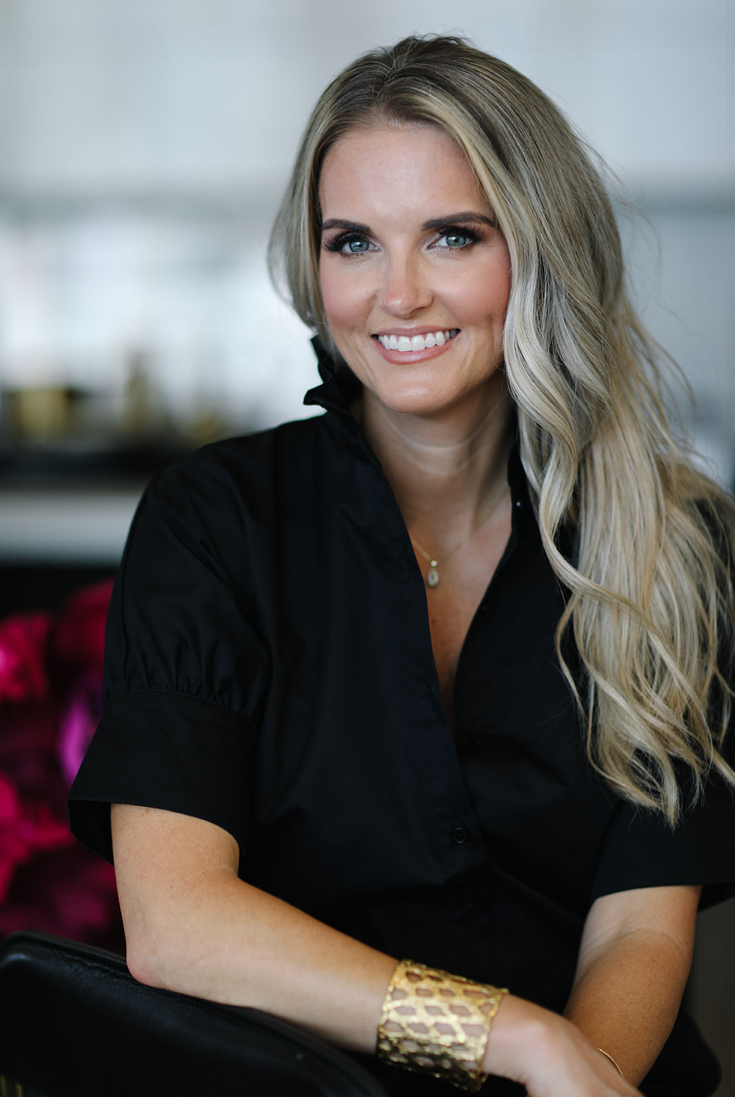Woman with blonde hair wearing a black top and gold bracelet indoors.