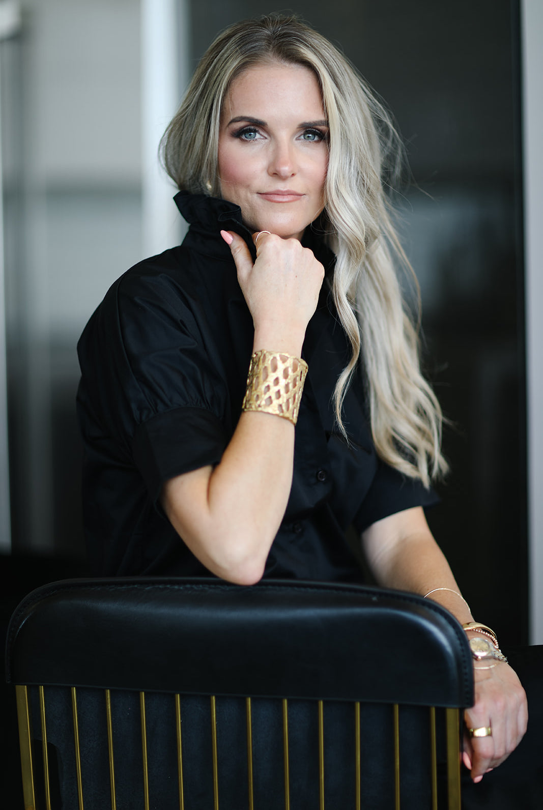 Woman with blonde hair and gold bracelet sitting on a black chair.