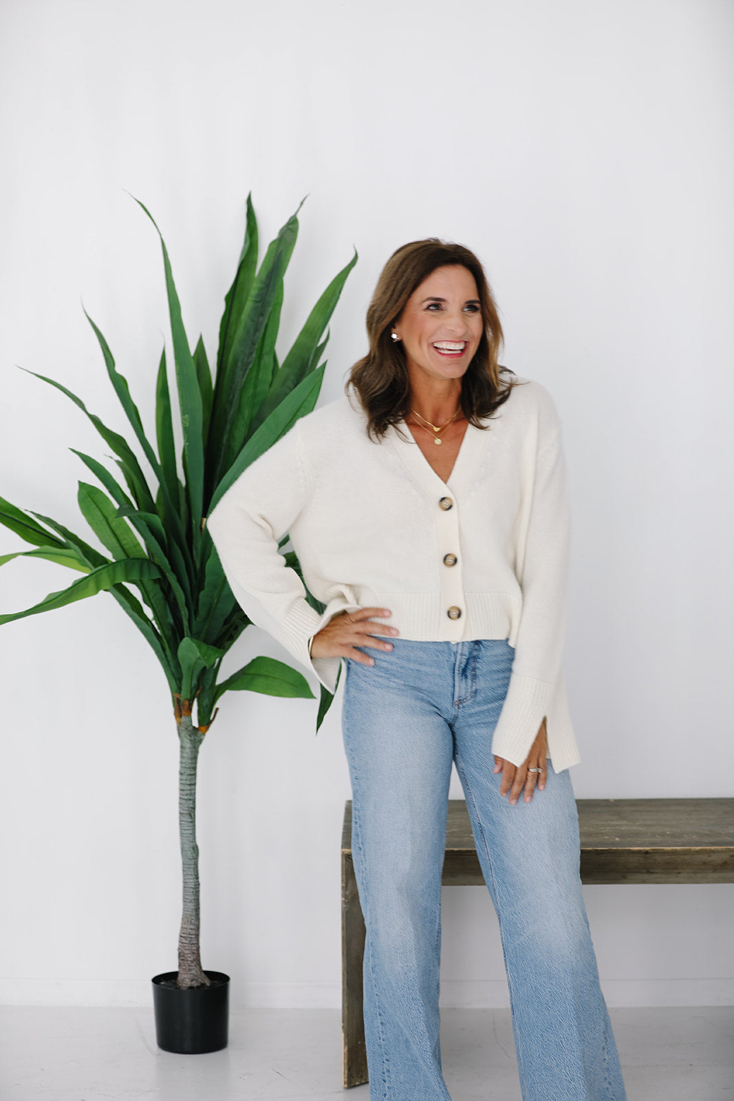 Woman in a white cardigan and blue jeans standing next to a plant against a white wall.