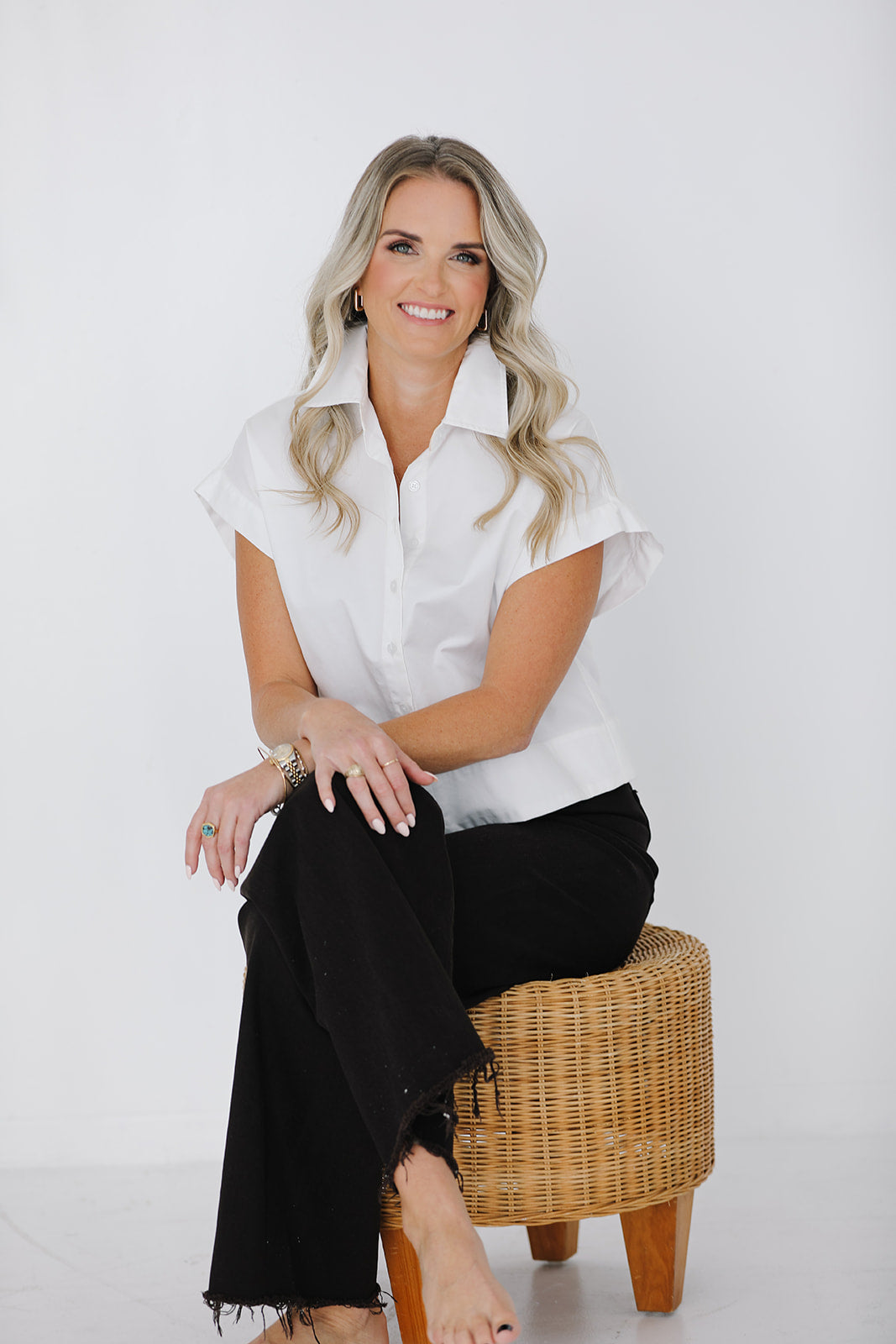 Woman sitting on a wicker stool wearing a white blouse and black pants against a white background