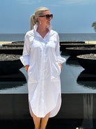 Woman wearing a crisp white shirt dress. The dress has pockets and has a mid length.