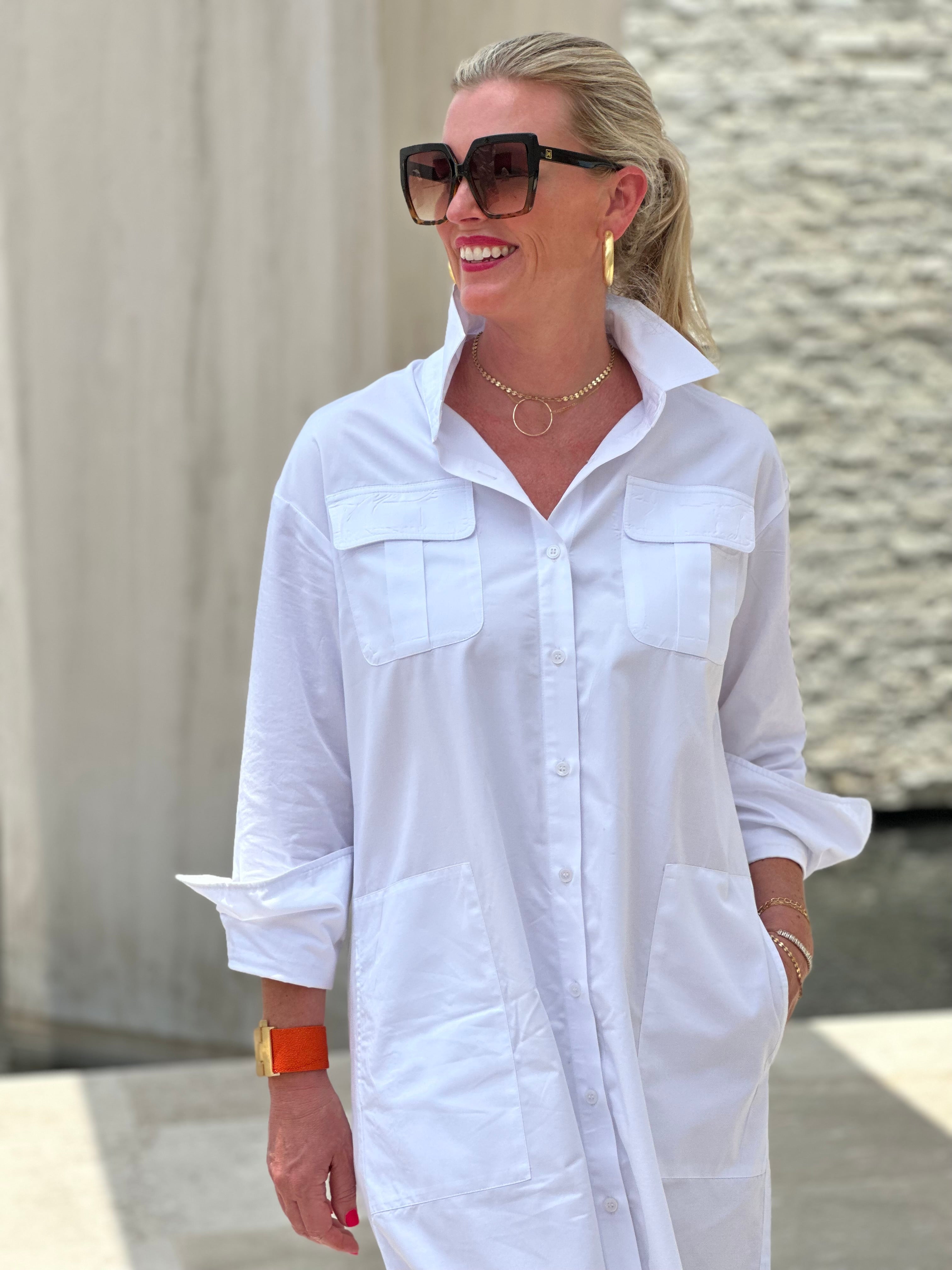 Woman wearing a crisp white shirt dress. The dress has pockets and has a mid length.