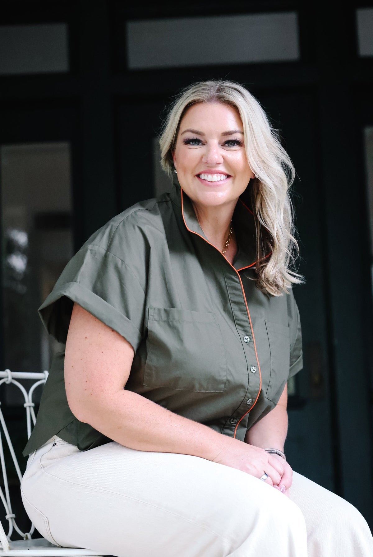 A smiling blonde woman sits on a white chair wearing an olive green short-sleeve button-up shirt with thin orange piping and white pants, posed in front of a dark doorway.