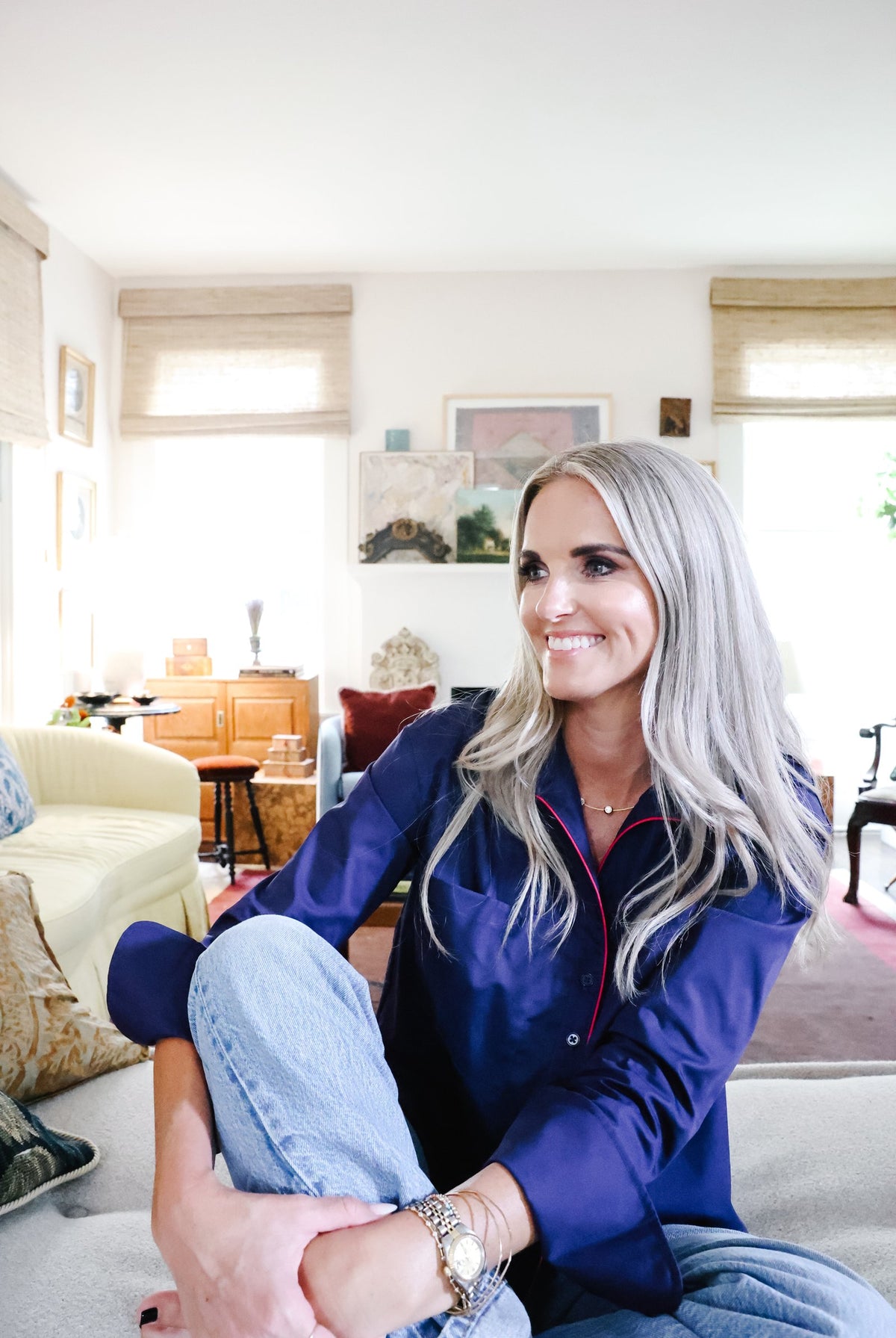 A smiling woman with long silver hair sits cross-legged on a light-colored sofa in a bright living room. She is wearing a dark navy blue satin pajama shirt with red piping and light blue jeans.