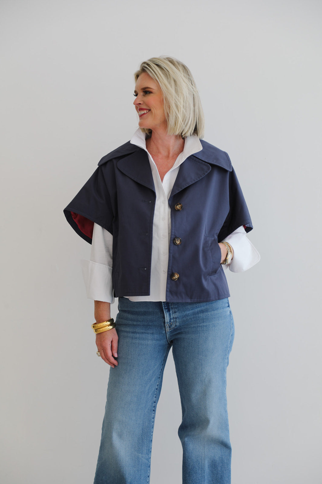 Woman wearing a navy jacket over a white shirt with rolled-up sleeves, paired with blue jeans on a plain background.