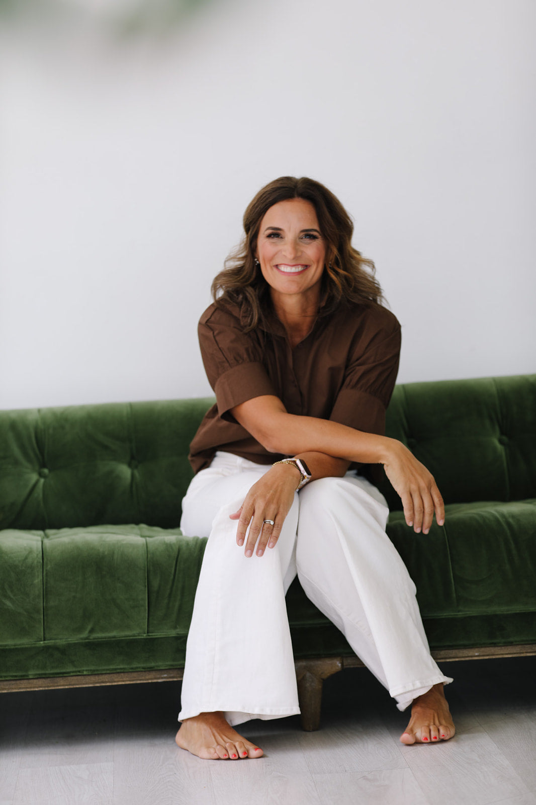 Woman sitting on a green couch wearing a brown shirt and white pants.