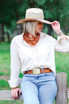 A woman wearing a straw cowboy hat, cream button-up shirt, brown belt, and light blue jeans, sitting on a wooden fence in an outdoor park setting.