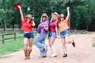 Four women in cowboy hats and boots posing on a dirt path with trees in the background