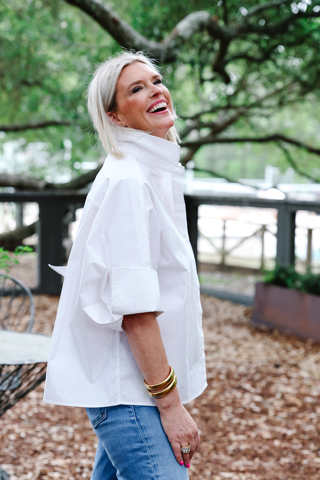 A woman with blonde hair wearing a white oversized mock-neck shirt and blue jeans, standing outdoors in a park-like setting with trees and a fence in the background.