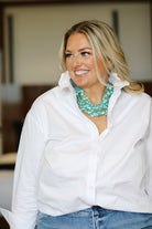 Woman wearing a white ruffle collared shirt and blue jeans with a green necklace indoors.