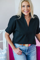 A smiling blonde woman wearing a black short-sleeve collared blouse tucked into light-wash blue jeans with a brown belt, standing indoors against a light wall.