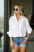 Woman wearing a white blouse with red accents and denim shorts, standing indoors.
