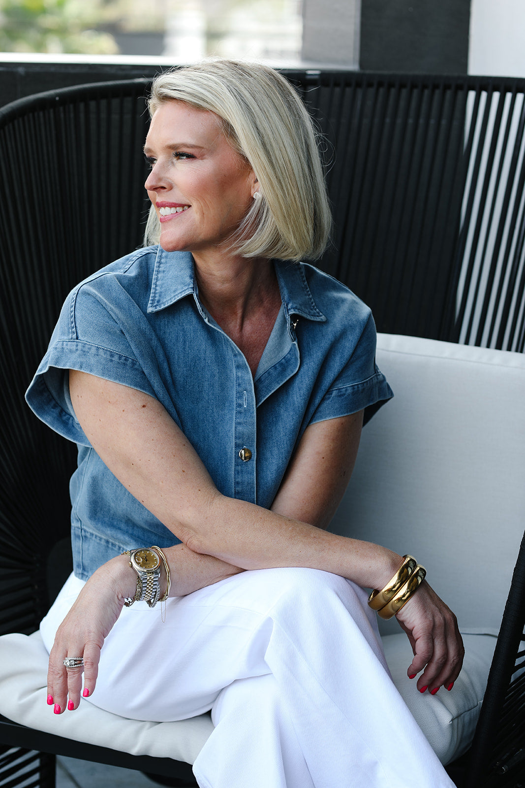 Woman sitting on a chair wearing a denim shirt and white pants, with a blurred background.