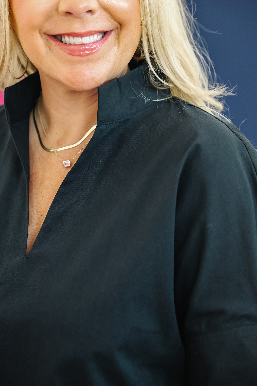 Close-up of a smiling blonde woman wearing a black V-neck top and layered gold necklaces, with a dark blue background.