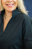 Close-up of a smiling blonde woman wearing a black V-neck top and layered gold necklaces, with a dark blue background.