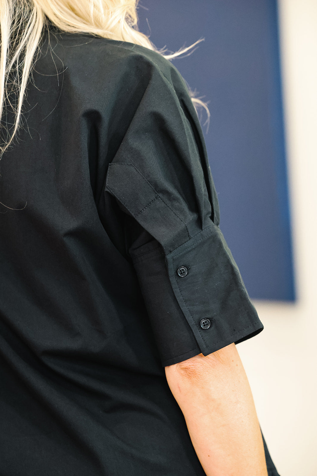Close-up of a person wearing a black short-sleeve top, showing the side and back of the sleeve with two buttons, against a blue and white background.