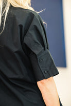 Close-up of a person wearing a black short-sleeve top, showing the side and back of the sleeve with two buttons, against a blue and white background.