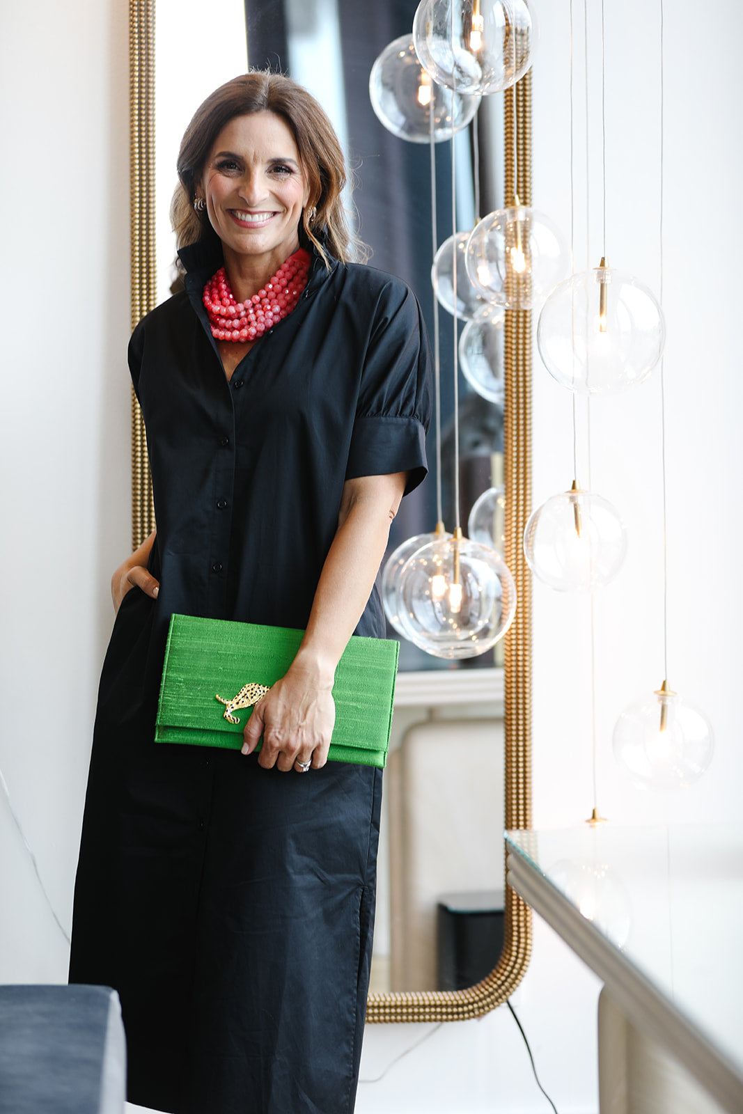 Woman in a black dress holding a green clutch in an indoor setting with modern decor.