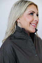 A smiling blonde woman shown in profile wearing a brown button-up shirt with a high ruffled collar and gold hoop earrings, against a plain light gray background.