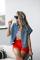 Woman in denim shirt, floral top, and red shorts holding a glass of wine indoors.