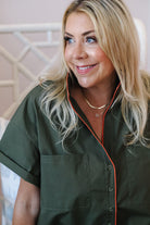 A smiling blonde woman wearing an olive green short-sleeve button-up shirt with orange piping trim, photographed in a softly lit indoor setting.