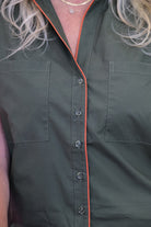 Close-up of a person wearing an olive green sleeveless button-up shirt with bright orange piping along the collar, placket, and pocket edges.