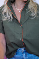 An olive green short-sleeve button-up shirt with bright orange piping trim along the collar and front placket, worn by a model with blonde hair and blue jeans.