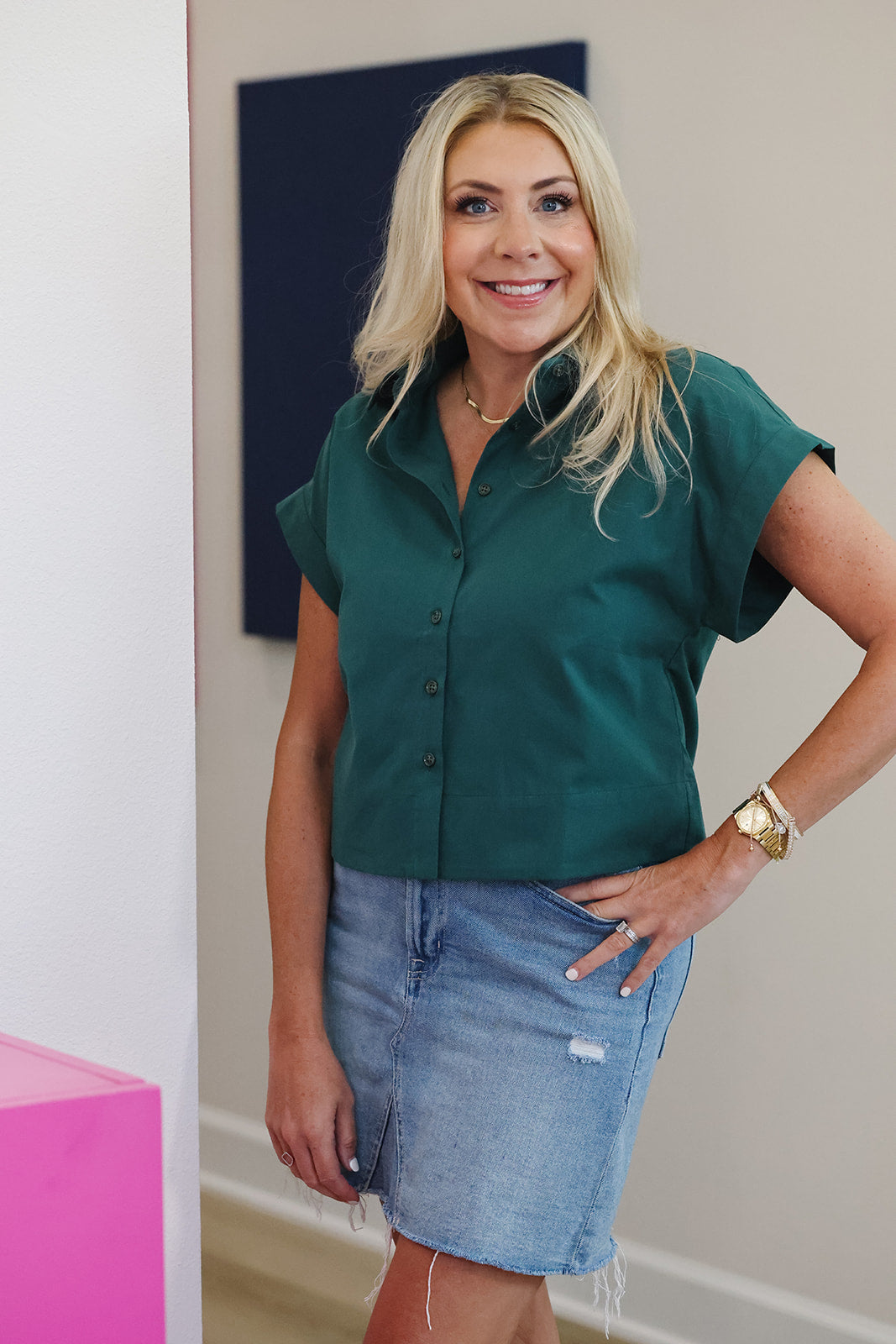 A woman with blonde hair wearing a dark green short-sleeve button-up shirt and a light blue denim skirt, standing in an indoor setting.