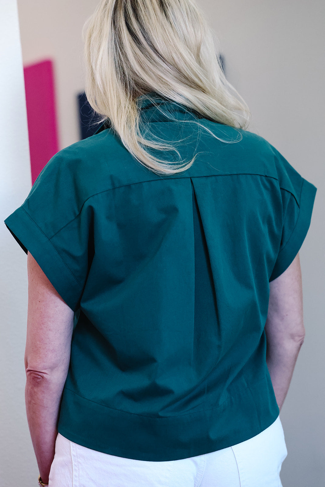 A woman viewed from behind wearing a dark teal green short-sleeve button-up shirt and white pants in an indoor setting.