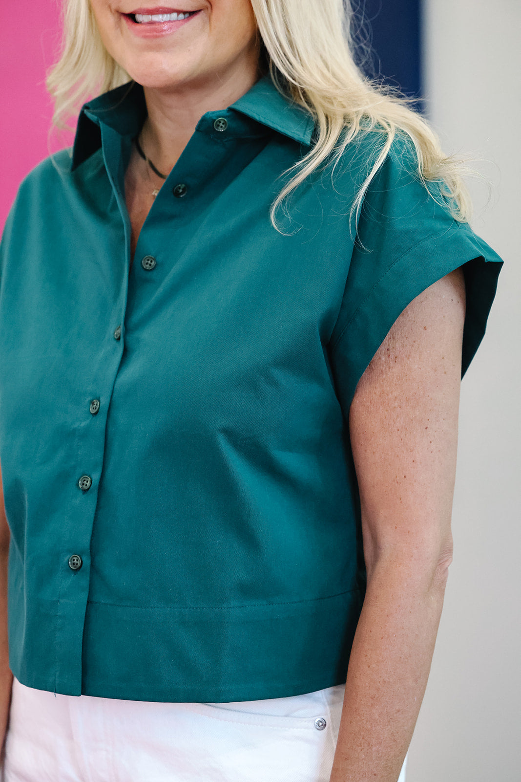 A woman wearing a teal green short-sleeve button-up shirt with white pants, shown from the chest up against a neutral indoor background.