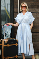 Woman in a light blue dress holding a suitcase and jeans, standing against a wooden wall.