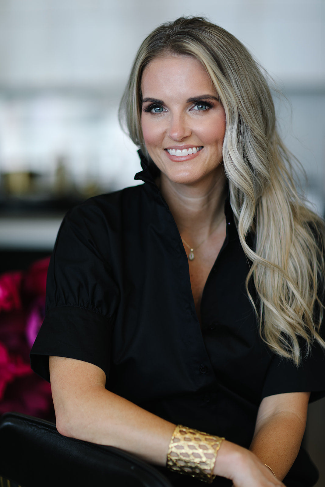 Woman with blonde hair wearing a black top and gold bracelet indoors.