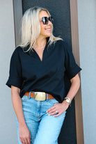 A woman wearing a black short-sleeve blouse tucked into light blue jeans with a brown belt and gold buckle, standing against a gray wall.
