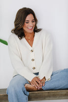 Woman wearing a white cardigan and blue jeans sitting on a wooden bench against a white wall.