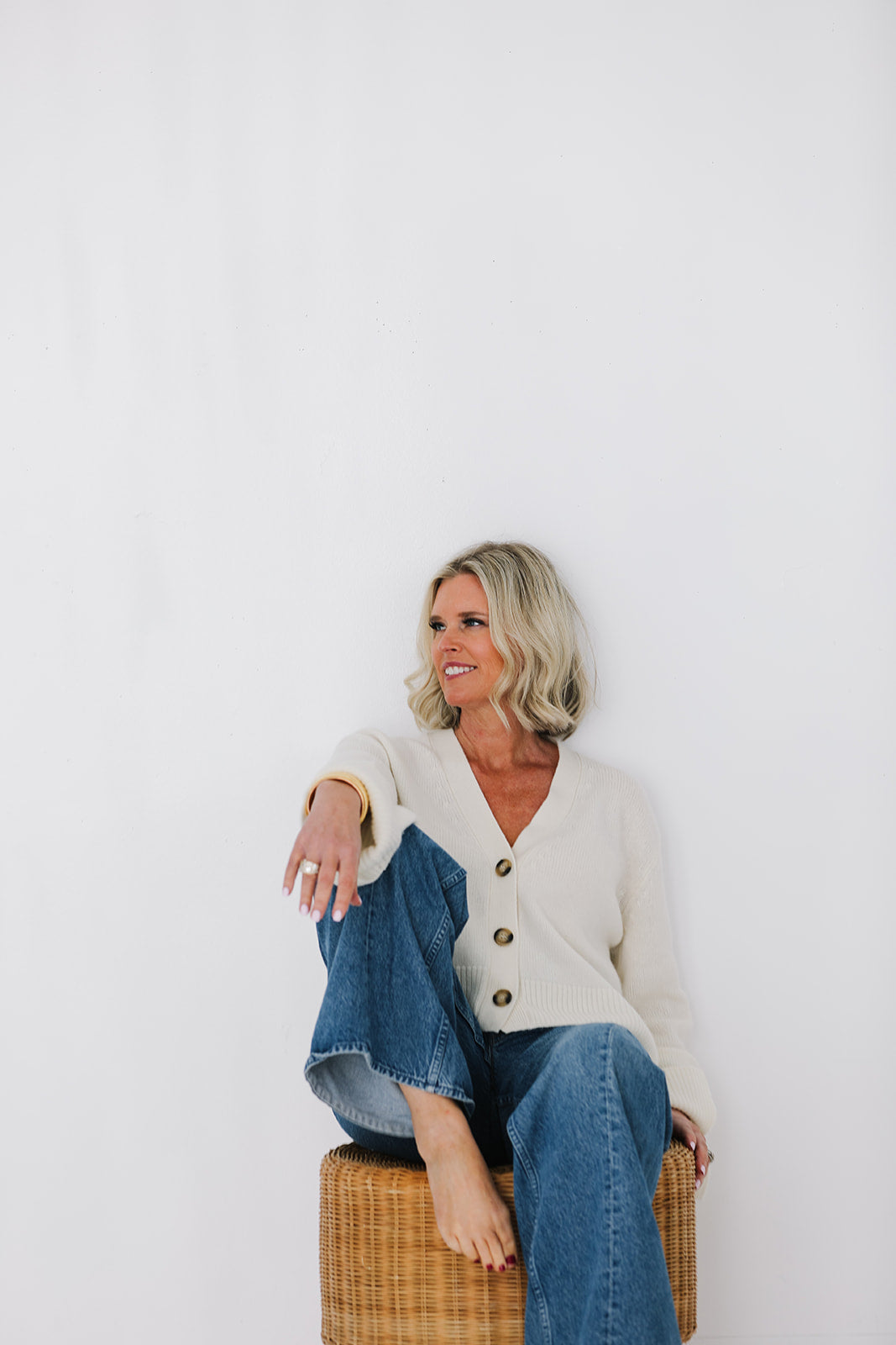 Woman sitting on a wicker stool wearing a white cardigan and blue jeans against a white background
