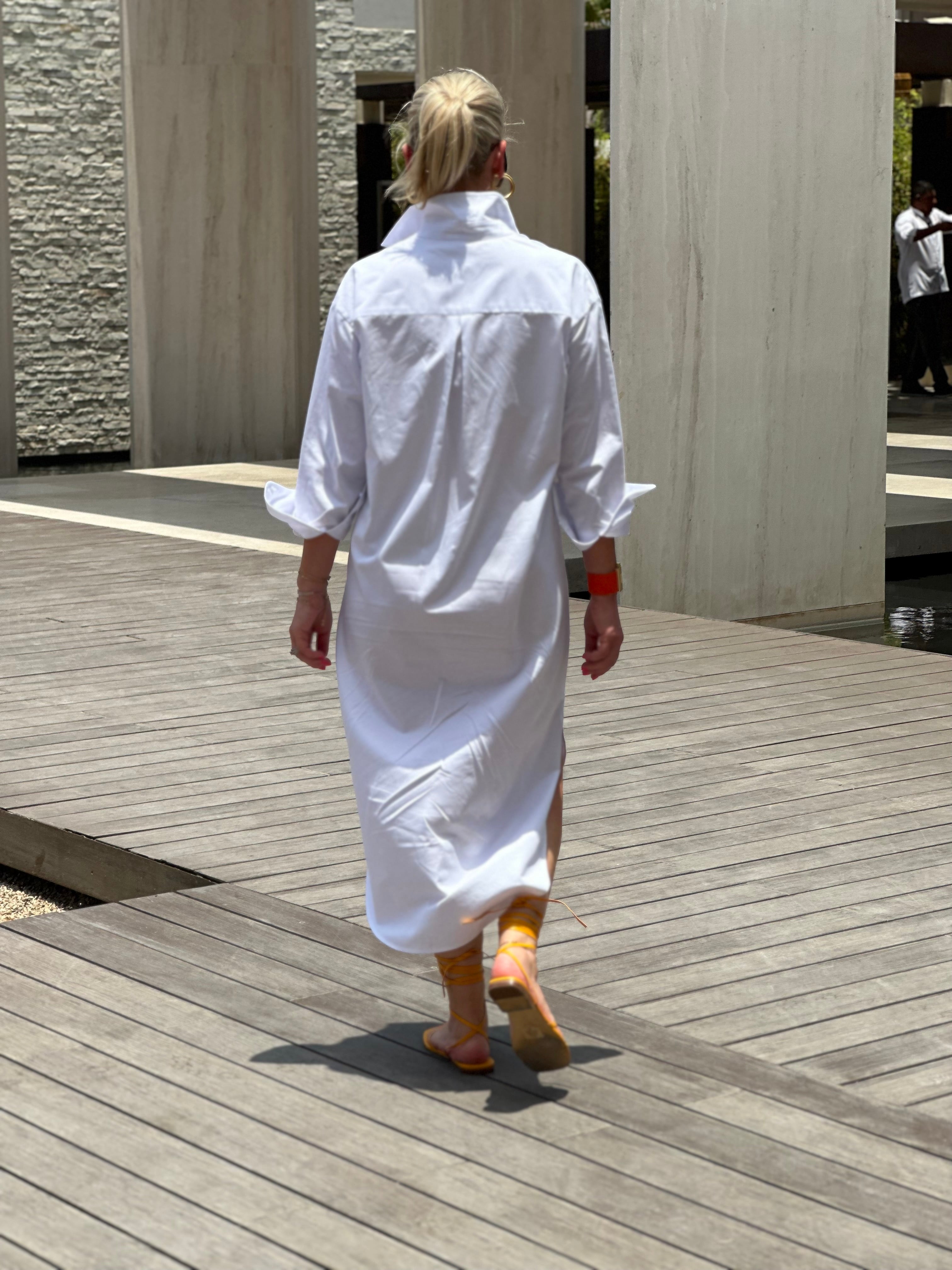 Woman wearing a crisp white shirt dress. The dress has pockets and has a mid length.