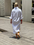 Woman wearing a crisp white shirt dress. The dress has pockets and has a mid length.