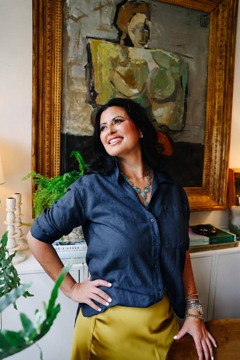 A woman posing in a navy blue button-down shirt and a mustard yellow skirt, standing in front of a large framed painting in an indoor setting.